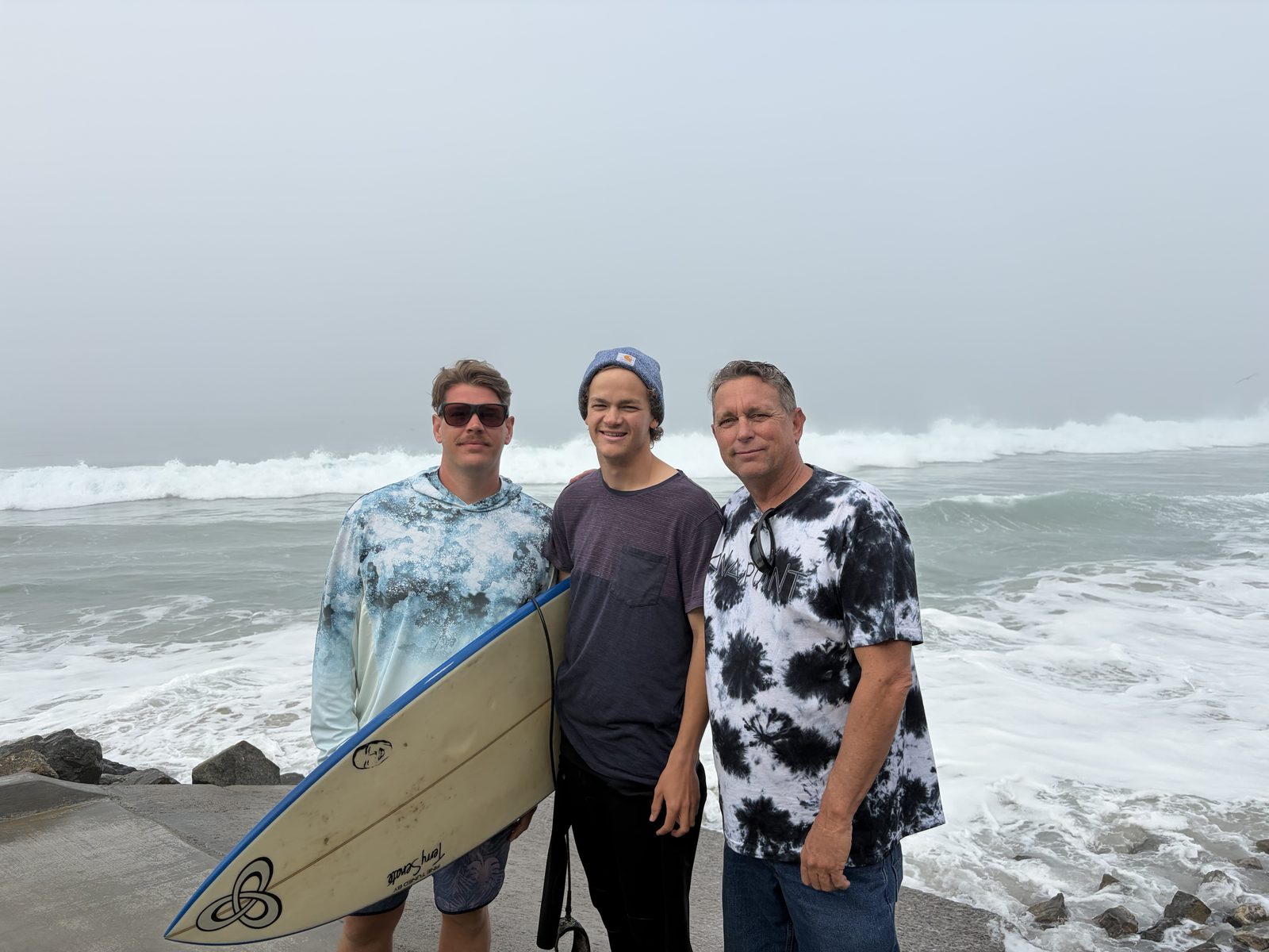 Three of the band on a fog-grey beach, one holding a surfboard, waves breaking behind them.