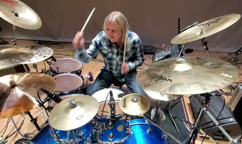 A drummer mid-strike behind a blue sparkle kit, sticks raised, cymbals crowding in from every edge of the frame.