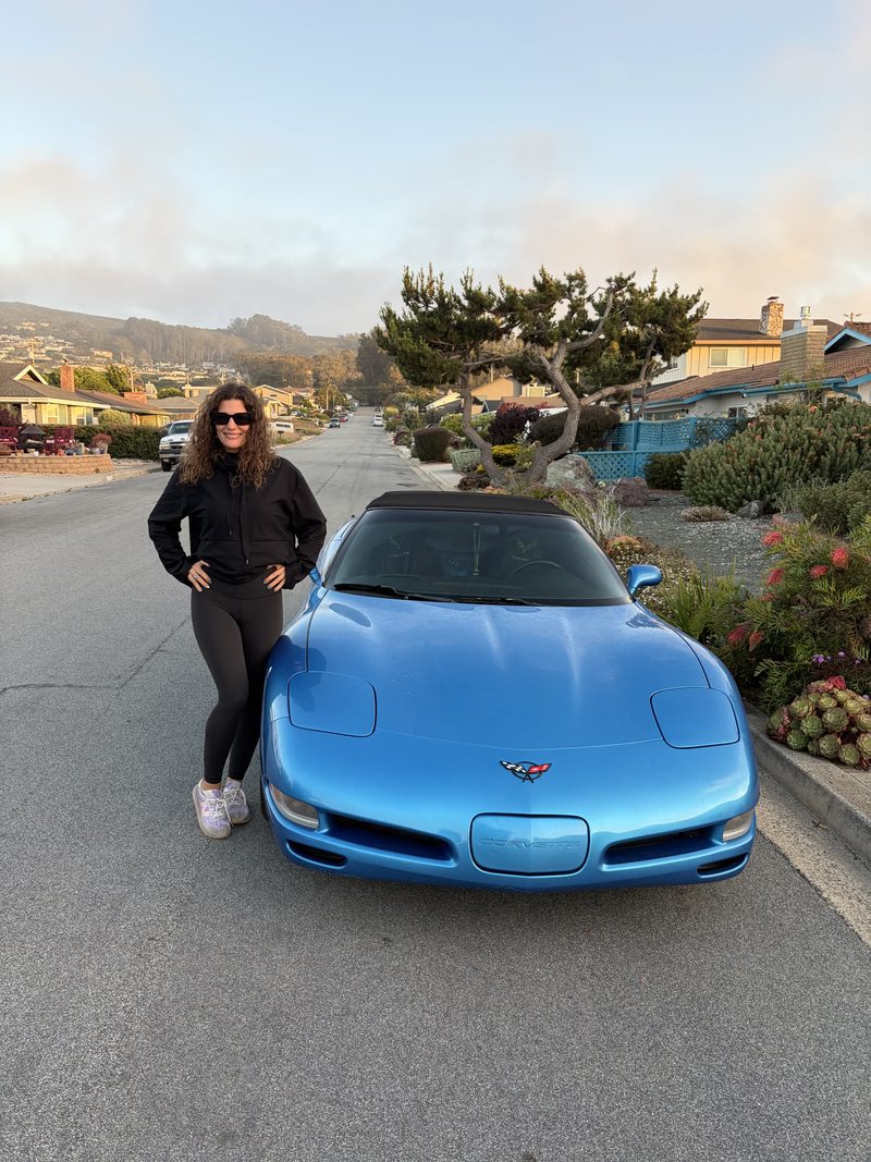 A woman in black leaning against the nose of a light blue Corvette convertible on a hillside street, fog sitting over the town behind her.