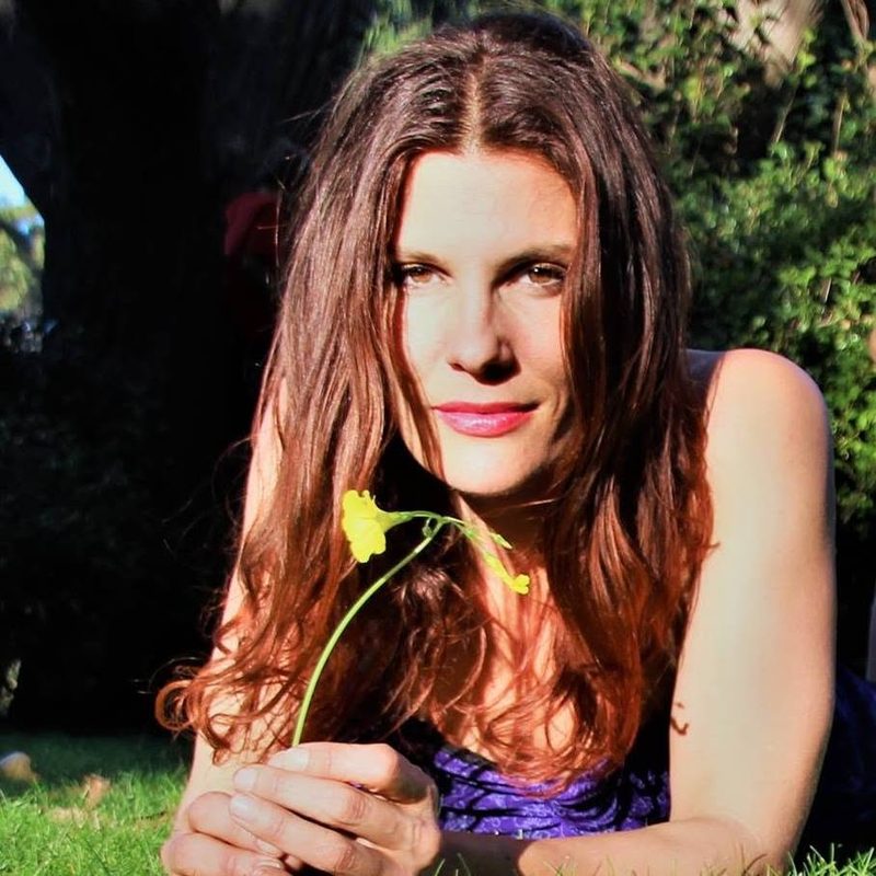 A woman lying in the grass in a purple top, holding a yellow flower out toward the camera.