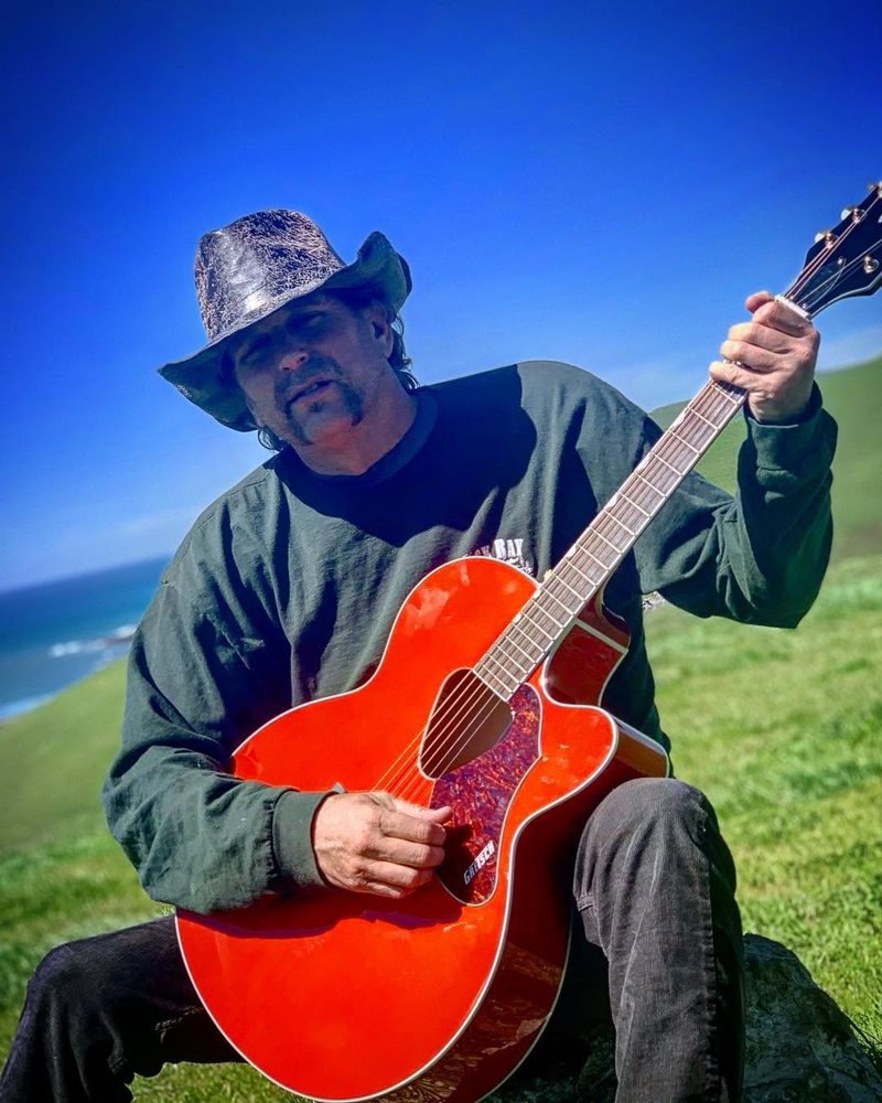 A man in a wide-brimmed hat playing a red archtop guitar on a grassy bluff above the ocean.