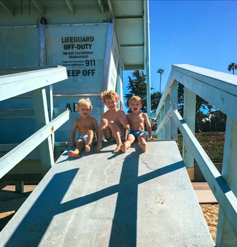Three small kids sitting in a row on the ramp of a lifeguard tower, palm trees and hard sun behind them.