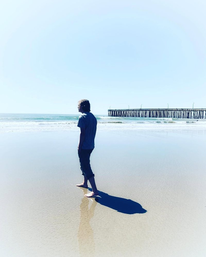 A lone figure standing on wet sand at the shoreline, long shadow running toward a pier in the distance.