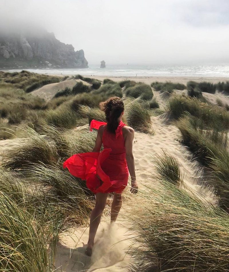 A woman in a red dress walking barefoot through beach grass and dunes toward the surf, Morro Rock in the mist beyond.