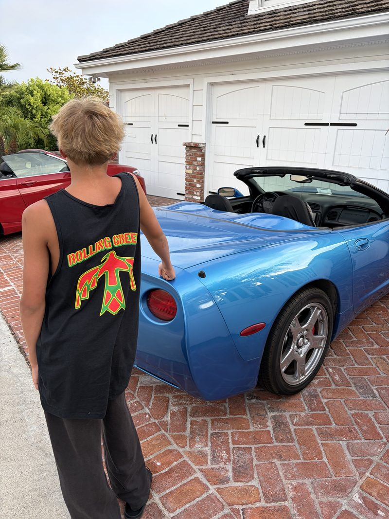 A kid in a black Rolling Greens thunderbird tank top walking toward a blue convertible in a driveway.