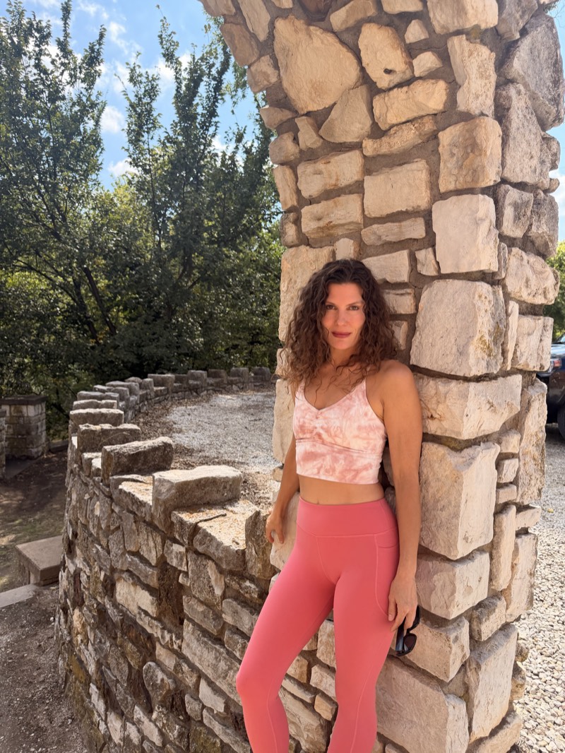 A woman with long curly hair in a pink top and leggings leaning against a stone pillar beside a low curved stone wall, trees behind her.