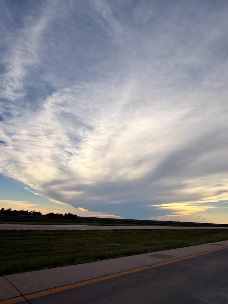 Streaked cirrus clouds burning orange over a flat coastal horizon at dusk, the edge of a road in the foreground.